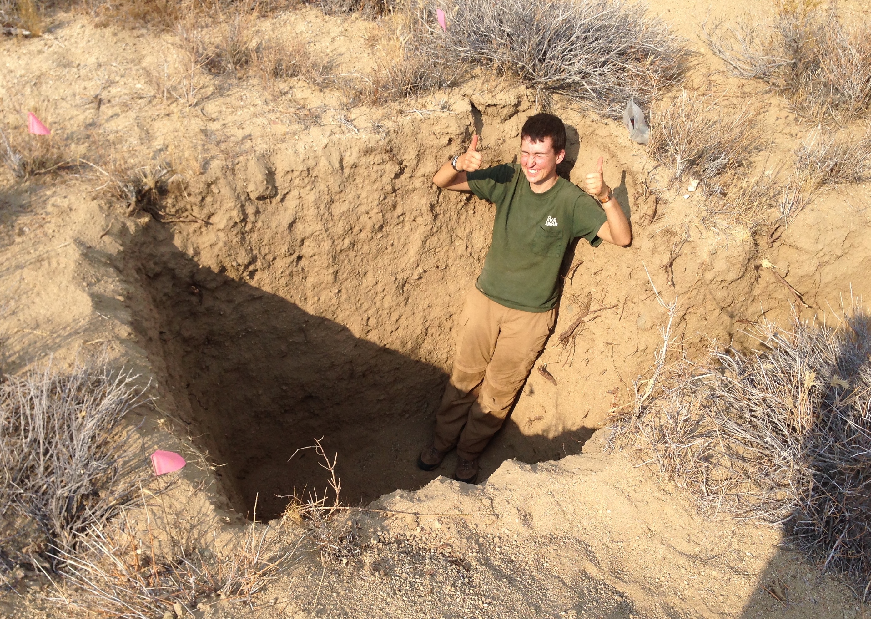white person standing in a soil pit (in semi-arid environment) qearing field clothes gives two thumbs up
